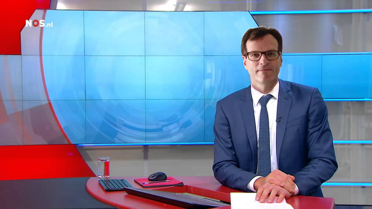 Rijzige man met rood-bruin haar in blauw pak achter een tafel met de handen over elkaar op een vel wit papier, tegen een decor van een nieuwsuitzending van omroep NOS.